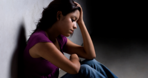 Young woman sits against a wall with her head resting on one hand, looking distressed or sadbedful.