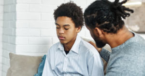 Young person in a light blue shirt sits with eyes down while a second person places a reassuring hand on their shoulder on a couch in a living room.